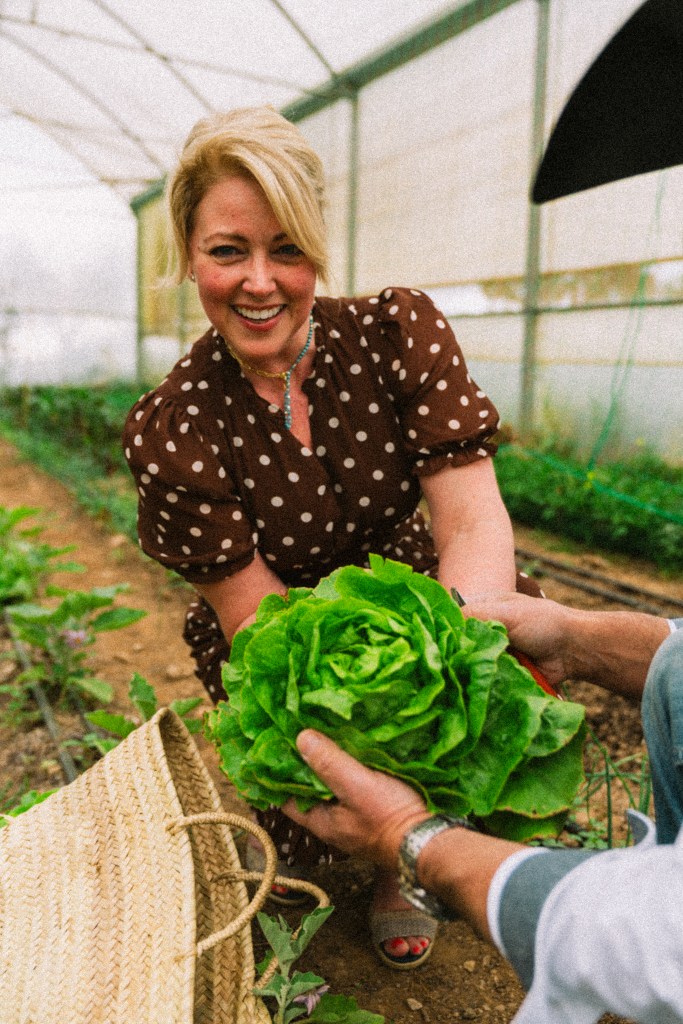 A woman wearing a polka dot dress joyfully hands over a large head of lettuce to another person in a greenhouse filled with greenery.