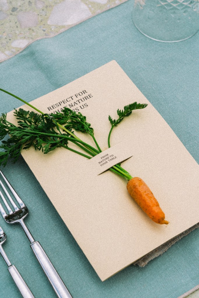 A menu featuring a small carrot with green leaves, placed on a beige background with the text 'RESPECT FOR NATURE' and 'FROM NATURE TO YOUR TABLE' visible, laid on a light blue tablecloth next to silverware.