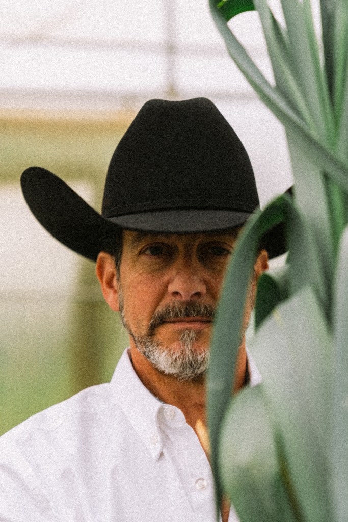 A man wearing a black cowboy hat stands partially obscured by green plant leaves, gazing directly at the camera with a serious expression.