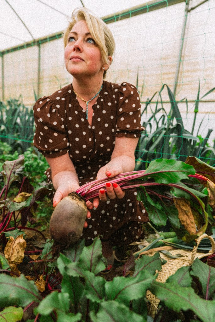 A woman in a polka dot dress holds a beetroot while surrounded by lush green plants in a greenhouse.