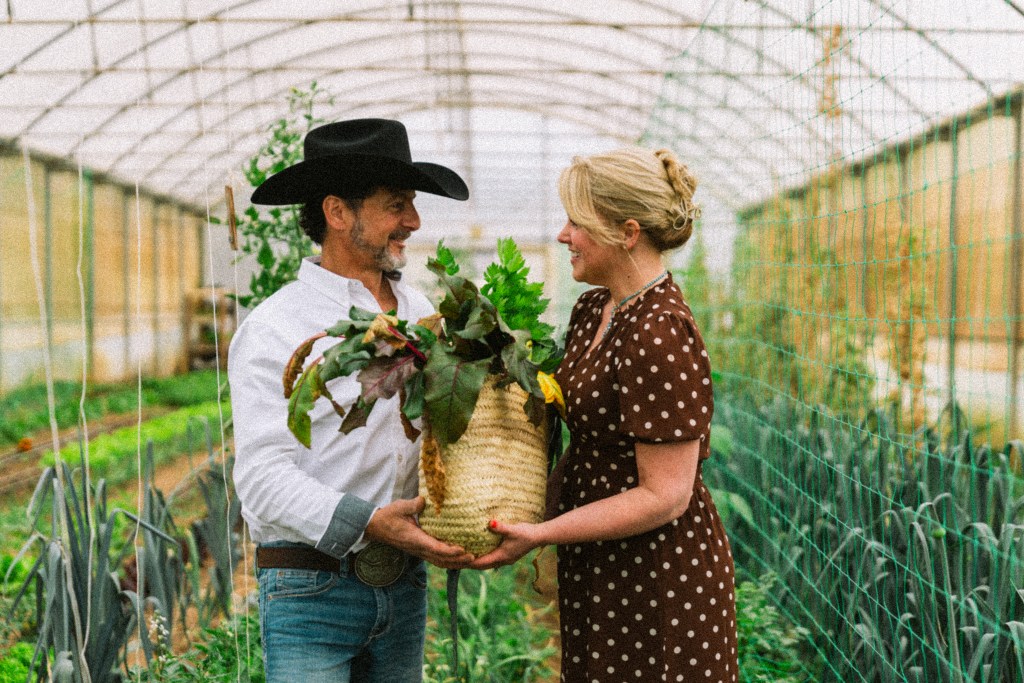 A smiling couple in a greenhouse, holding a basket of freshly harvested vegetables. The man wears a black cowboy hat and a white shirt, while the woman is dressed in a brown polka dot dress.