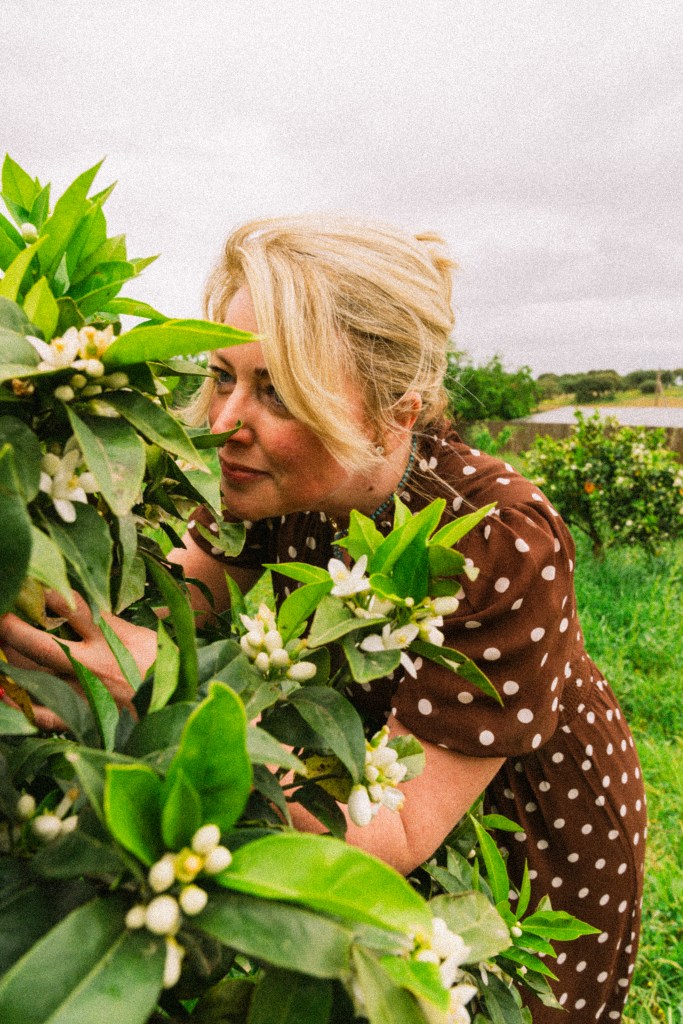 Woman with blonde hair in a brown polka dot dress tending to a flowering citrus tree in a green field.