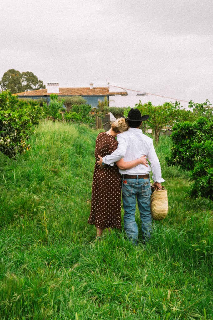 A couple standing together in a lush green field, with the woman dressed in a polka dot dress and the man in a white shirt and cowboy hat. They are embracing and facing towards a house in the background.