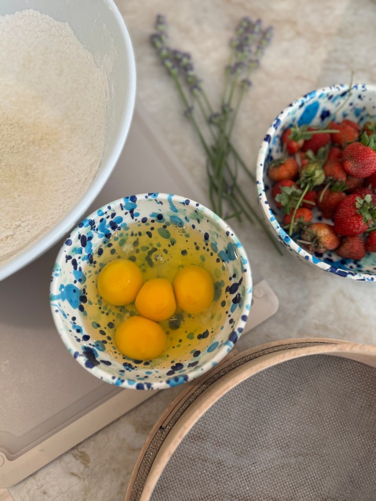 Ingredients for baking including a bowl of eggs, a bowl of strawberries, flour, and sprigs of lavender.