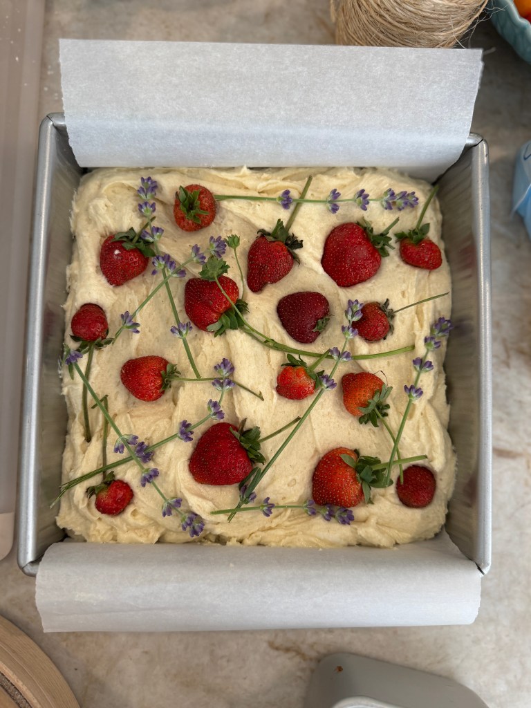 A rectangular cake in a metal baking pan, topped with fresh strawberries and sprigs of lavender on a creamy frosting.