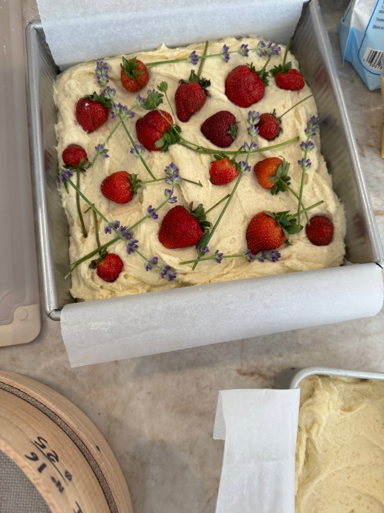 A square cake batter topped with fresh strawberries and sprigs of lavender, placed in a baking pan lined with parchment paper.