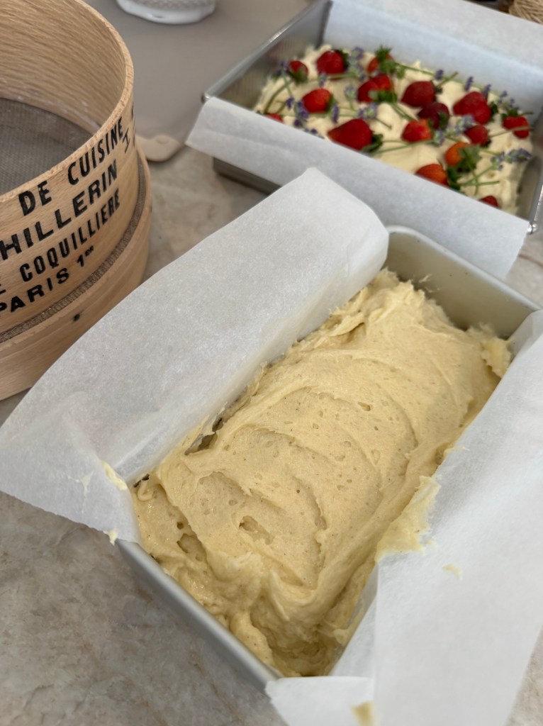 A baking dish filled with yellow cake batter, surrounded by a wooden sieve and another dish topped with strawberries and flowers.