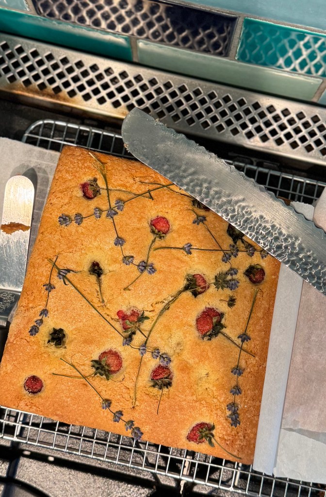 A rectangular cake decorated with dried flowers and herbs, including lavender and rose petals, resting on a wire cooling rack with a metal spatula next to it.