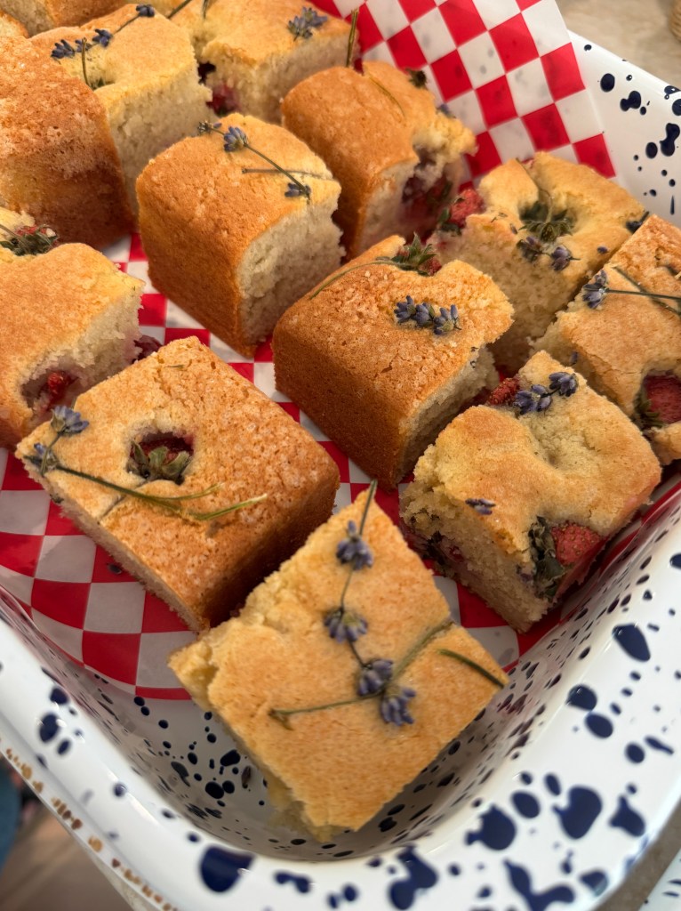 A tray of square pieces of cake decorated with lavender and strawberries, presented in a dish lined with red and white checkered paper.