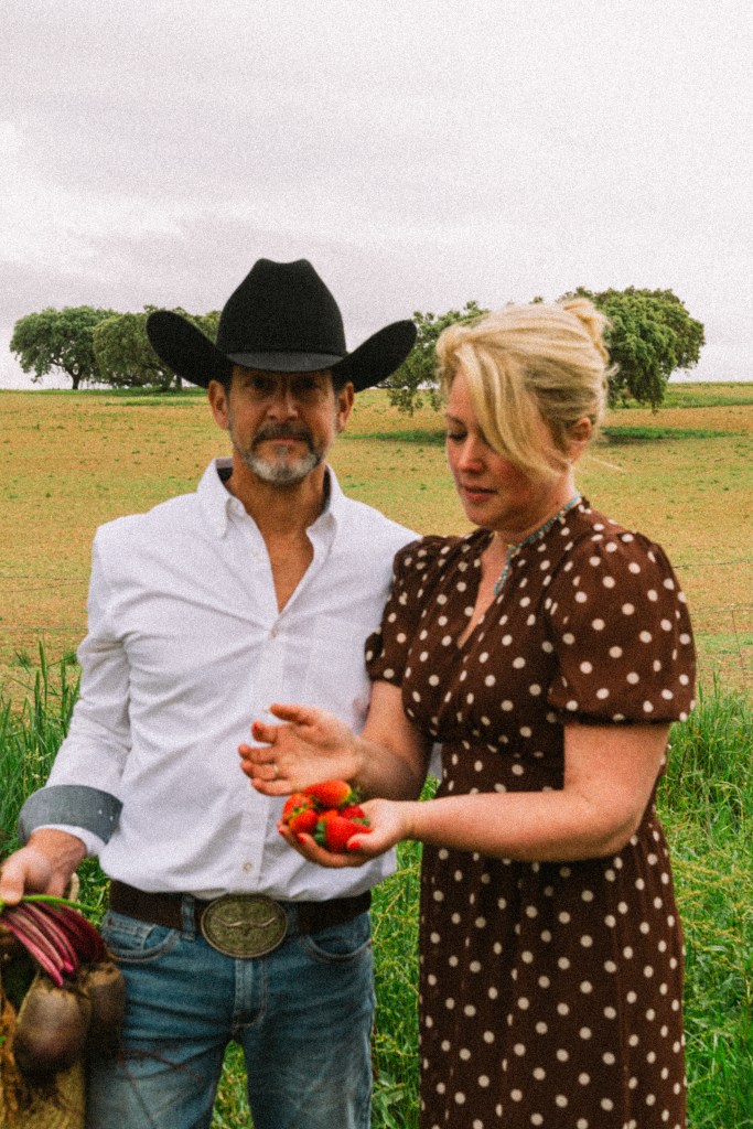 A man in a white shirt and cowboy hat stands beside a woman in a brown polka dot dress. The woman holds strawberries in her hands while the man carries vegetables in a bag, set against a green field.